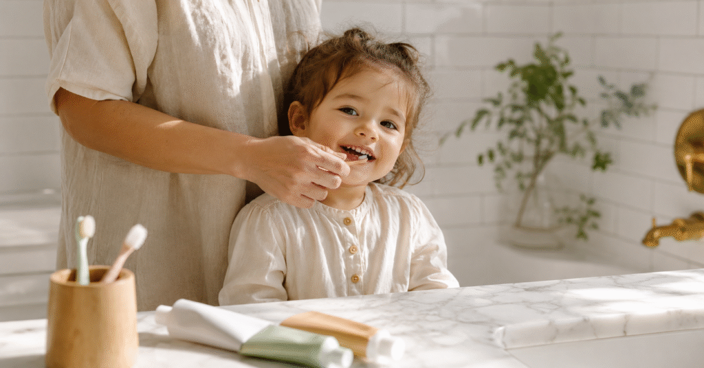 A toddler smiling while holding a toothbrush, with mom standing behind in a bright white bathroom — illustrating a guide to the best toddler toothpaste