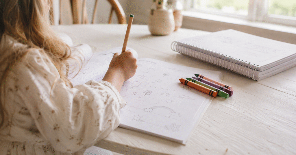 Open preschool activity workbook with a child's hand holding a pencil and crayons on a bright white table