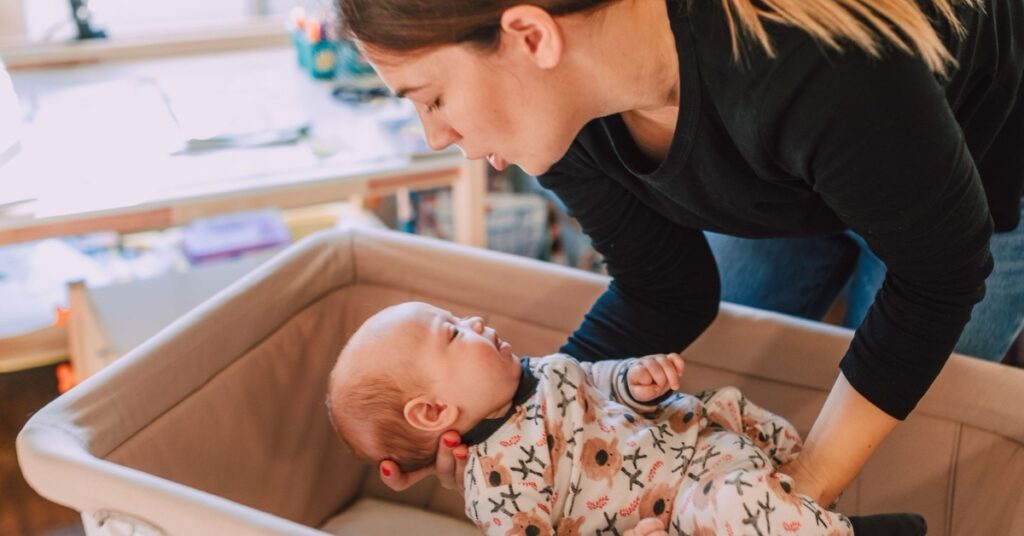 A mother supports her awake baby above an empty bassinet in a warmly lit nursery.