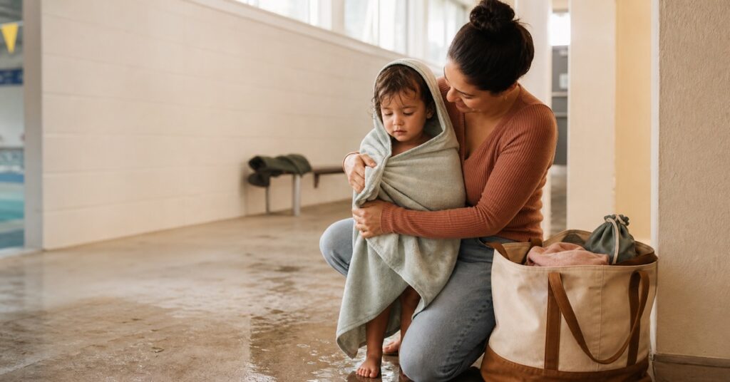 Parent wrapping a toddler in a hooded towel beside an organized swim bag after an indoor swim lesson.